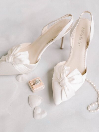 Wedding details shot of a white satin wedding shoes with a pink ring box and pearl necklace.