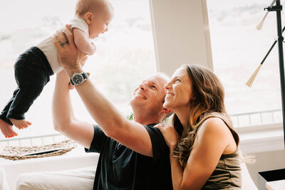 A father lifting his baby boy up in the air as mama looks on and smiles.