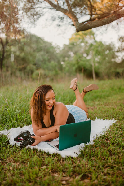 A female photographer works outdoors on a laptop and camera during a personal branding photoshoot at Challenger 7 Park in Friendship, Texas. She lies on a blanket in the grass, surrounded by nature.