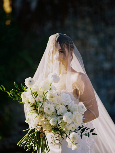 Bride holding a lush white rose bouquet beneath her veil, captured in soft golden light by Asia Pimentel Photography.