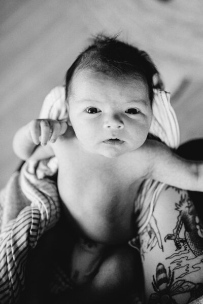 Candid black and white newborn portrait capturing a wide-eyed baby wrapped in a cozy blanket with natural light and soft textures.