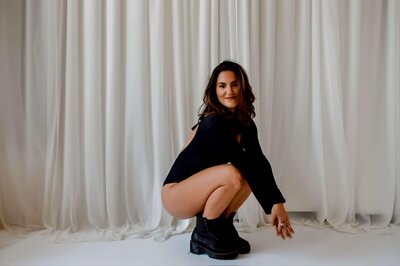 Emily Michele crouching confidently in a black bodysuit, smiling softly — representing feminine power, confidence, and embodiment for the Unveiled program.