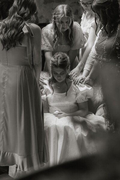 A bride is seated with her bridesmaids laying hands on her to pray over her.