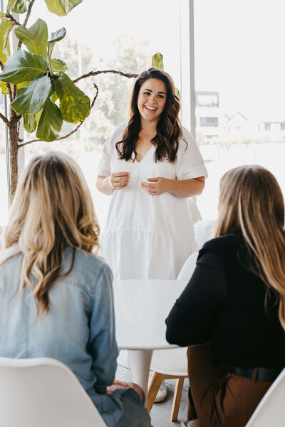 3 women talking, dark haired woman standing and teaching 2 blonde hair women sitting down