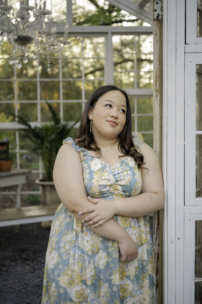 Beautiful high school senior wearing a blue and yellow floral dress leans against the doorway of a greenhouse
