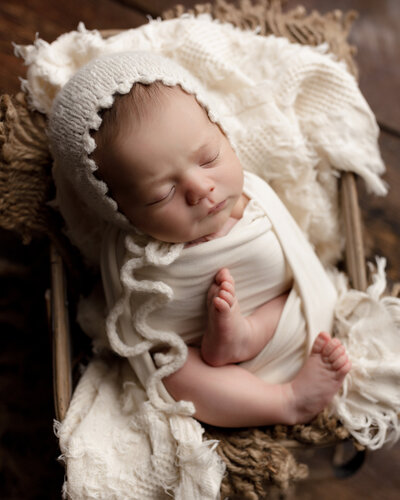 Newborn studio portrait by top newborn photographer. Baby laying in a basket and wrapped in a scream swaddle. Baby is wearing a cream coloured bonnet and sleeping. Baby's toes are peeking out of the swaddle.