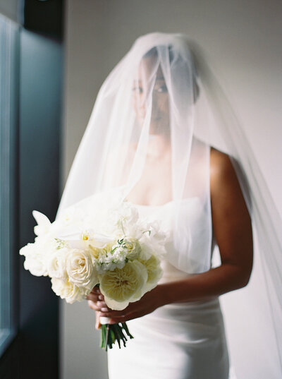 Film photograph of a Bride holding her bouquet under her veil as she stands next to a window.