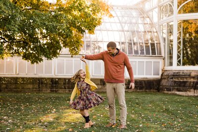 A young girl holding hands with an adult male while spinning around, both surrounded by autumn leaves with a greenhouse structure in the background. Photo captured by a family photographer Pittsburgh PA.
