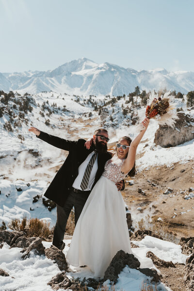 bride and groom holding each other in fun pose smiling wide holding boquet in the air standing on a cliff in front of towering mountain above a valley