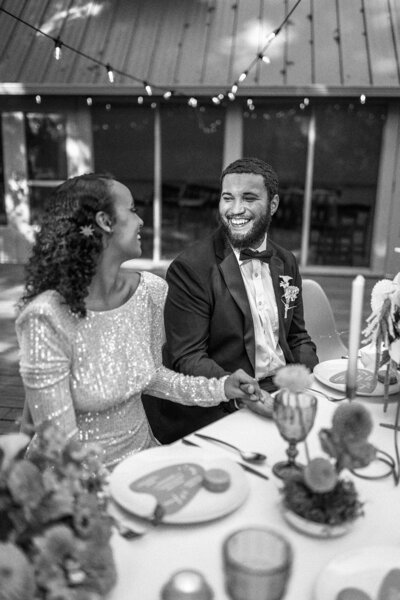 Bride and groom hold hands smiling each other at a table, representing an example of the documentary wedding photography style of Among the Oaks. 