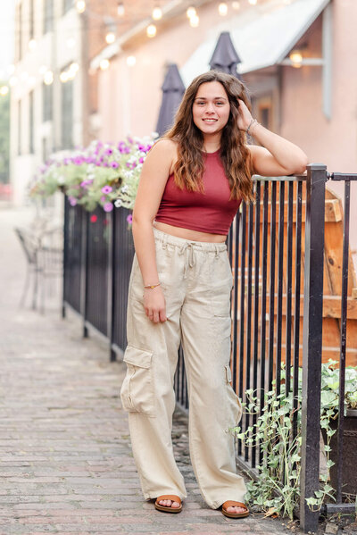 A high school senior, wearing a red crop top and cargo pants, leans on a black fence in an alley during her senior casual session.