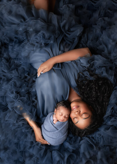 Newborn Photography -- Mother in flowing navy gown lies with her swaddled newborn baby on matching ruffled fabric in a Birmingham photography studio