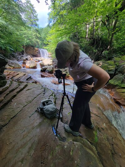 Joy Granados setting up her camera on a tripod by a waterfall, behind-the-scenes image of Capture Joy Photography, Northern Virginia family photographer.