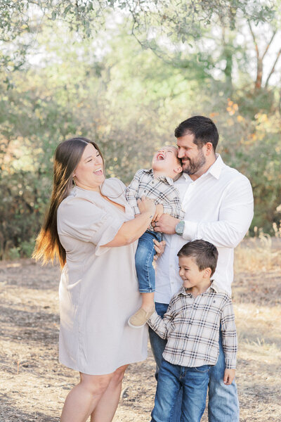 Family of four stands in front of a lake while smiling at the camera at Blue Hole Park, beautifully captured by an Austin Texas Portrait Photographer