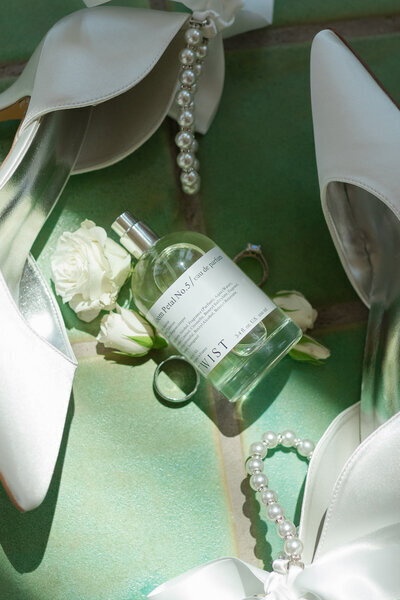 Elegant bridal shoes paired with pearls and floral perfume bottle on a shimmering green surface, highlighting sophisticated wedding details.