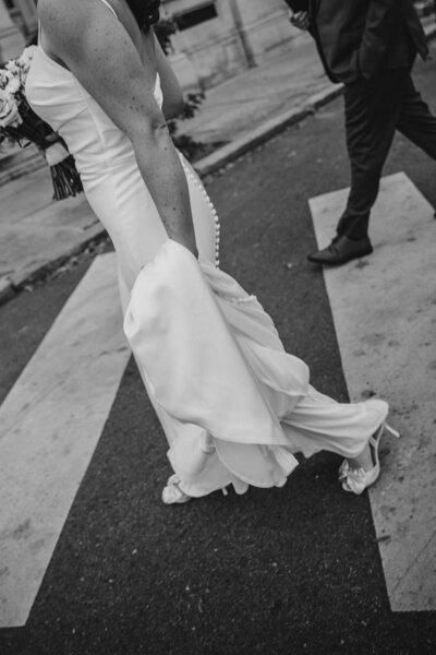 A newlywed couple walking across a crosswalk while one holds the bottom of their wedding dress 