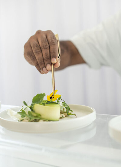 A chef's hand putting the final flower on a gourmet plate of food. The food is white and green and plates on a white plate.