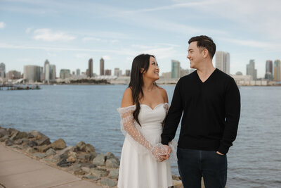 Smiling couple holding hands along the San Diego waterfront with the city skyline in the background.