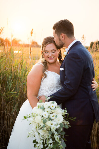 Warm golden light at sunset behind a bride and groom overlooking a marsh, the groom gently kisses the bride on the temple