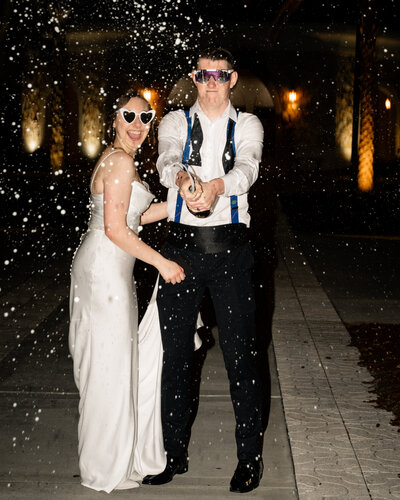 A Bride and groom do a champagne spray outside at night during their catholic wedding reception in St. Augustine, Florida. They are wearing sunglasses and laughing.