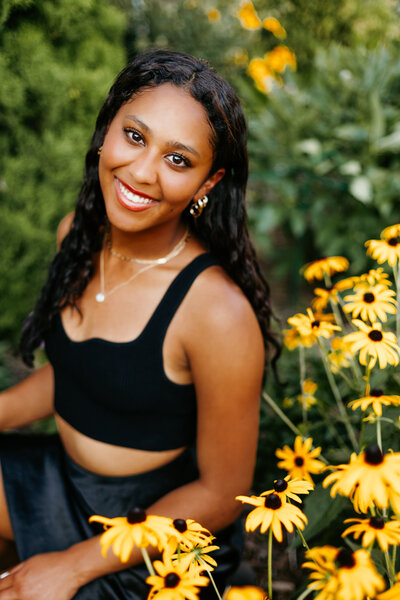 Senior girl in a black top posing in between green grass and yellow flowers