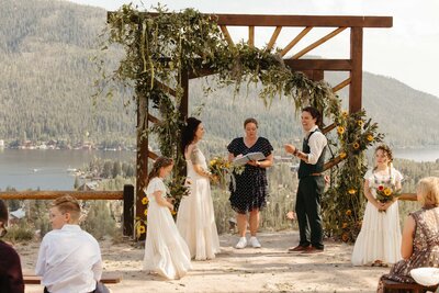 A wedding ceremony with a bride and groom in front of a wood arbor decorated with greenery with a lake and mountain view behind them