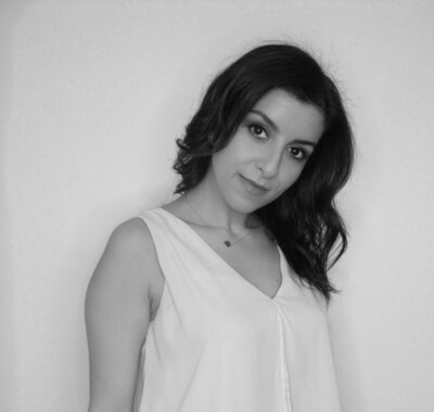 Black and white photo of woman with short brown, wavy hair and posing for the camera in a white sleeveless shirt.