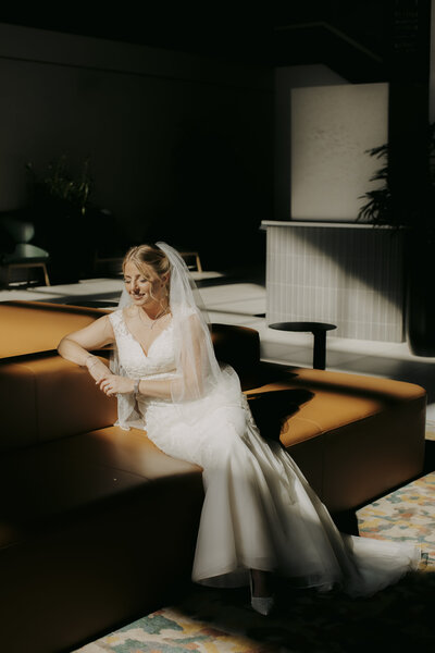 A bride in a lace gown and veil sits gracefully on a modern tan sofa, bathed in soft sunlight streaming through a window, creating a warm and serene atmosphere.
