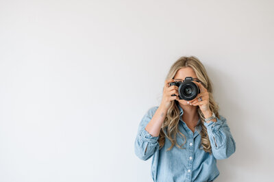 Emily sits on a clear plastic chair, looking straight ahead, not at the camera. She has her feet resting on the chair and hands on her knees, wearing light wash denim jeans and a matching denim button-up shirt.