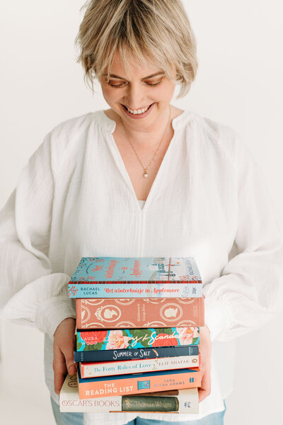 Rosanne van Cruijningen, zelfstandig redacteur en schrijfcoach voor romanschrijvers, houdt lachend een stapel boeken vast. 