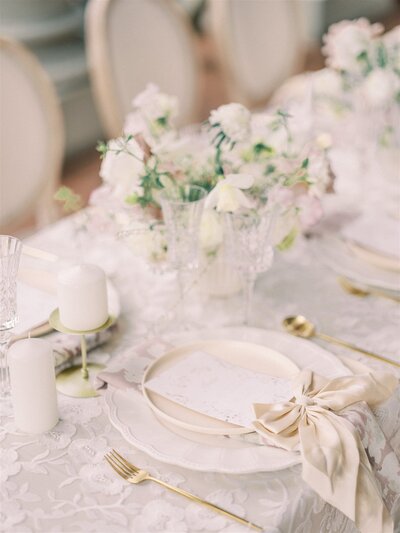 Elegant wedding table setting with a white lace tablecloth, white plates with a menu card tied by a beige ribbon, gold cutlery, white candles, and floral centerpieces featuring white and pastel flowers—perfectly curated by a top-notch Canadian wedding planner.