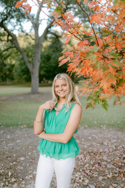 Georgia Southern graduating senior smiles at sweetheart circle next to the autumn colored tree. She is wearing a green top and white pants and there are trees in the background.