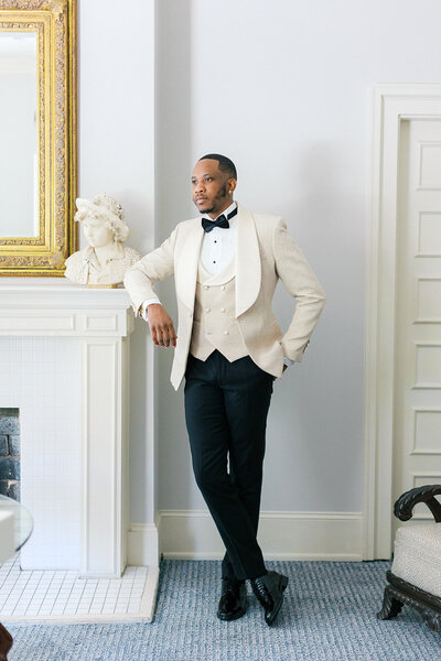 African American Groom leans against fireplace in white dinner jacket during wedding day at Wimbish House in Atlanta Georgia