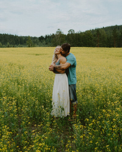 Man hugging his fiancé behind her back and she is smiling eyes closed in a field of rapeseeds in Finland