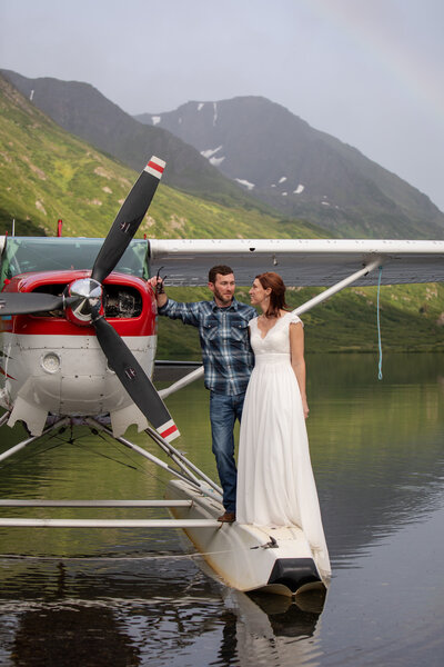 A bride in a white dress stands next to her groom in a flannel shirt on the float of a float plane in Alaska on their wedding day.