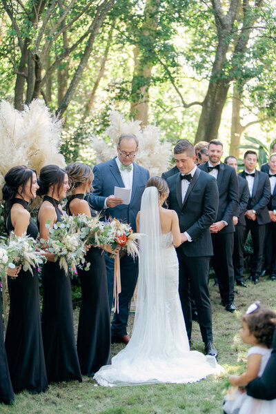 Golden light shines through outdoor garden wedding ceremony in Charleston. Bridesmaids in black.