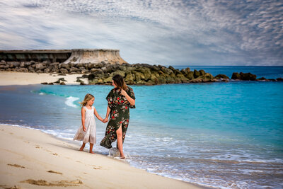 A mom holds hands with her daughter while they walk on the beach together.