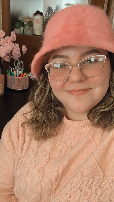 A woman, Rachel Manasco, wearing a pink fuzzy hat, pink-framed glasses, and a pink sweater. She is sitting at her desk where she works as a Showit Website Designer.