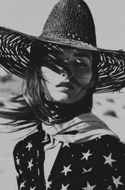 Black and white photo of a person in a star-patterned outfit and wide-brimmed straw hat, windblown hair veiling their face—a striking image styled by Sweet Pea Ink Creative, specialists in brand design.