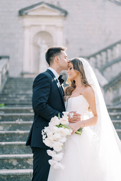 Groom kissing bride on the forehead while holding bouquet, gentle wedding portrait on historic staircase