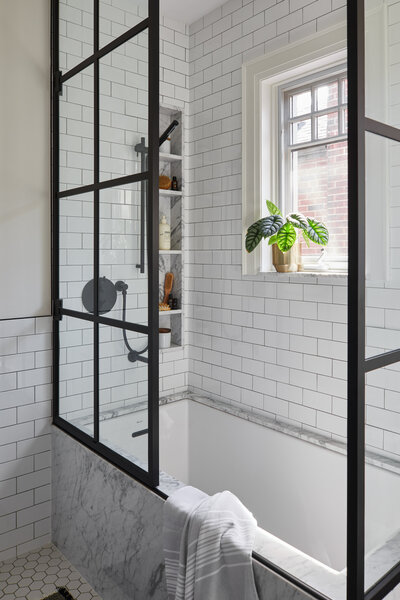 A rectangular marble bathtub surrounded by white subway wall tiles and a paned glass shower enclosure and a small window with a potted plant