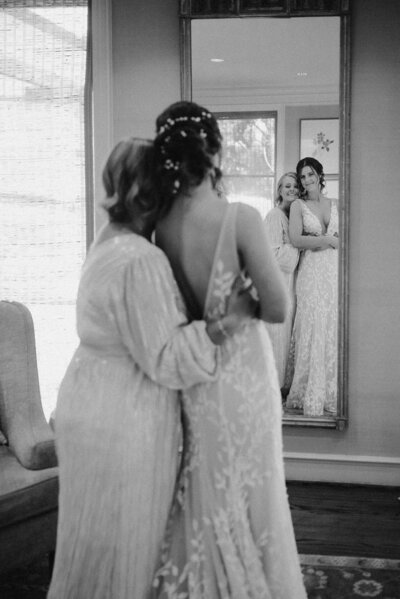 Black and white candid wedding photo of a bride embracing her mother while looking into a mirror, capturing an emotional getting-ready moment before the ceremony.
