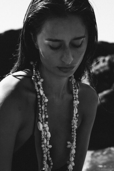 Black and white photo of the woman with wet hair, wearing layered shell necklaces, looking down thoughtfully against a rocky background. Captured by Sweet Pea Ink Creative, this image perfectly complements their distinctive brand identity.
