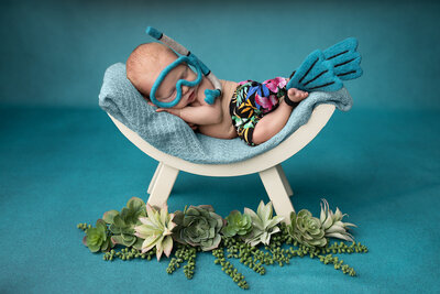 Newborn baby dressed in tropical swim trunks with felt snorkel gear and flippers, posed on a mini hammock surrounded by succulents, captured by Dallas newborn photography studio specializing in creative newborn pictures and baby photographer sessions.