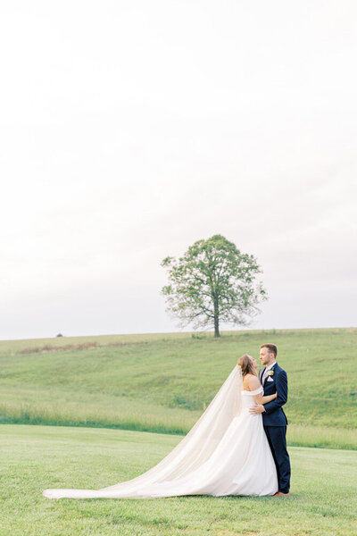 bride and groom embrace each other with long train and veil in green field