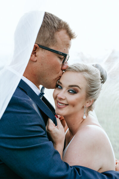 Bride holding onto groom’s jacket, staring into the camera while he kisses her gently on the temple.