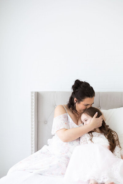 Mom kissing her daughter's head on white bed in Kathleen Jablonski's white studio  NH Maternity Photographer