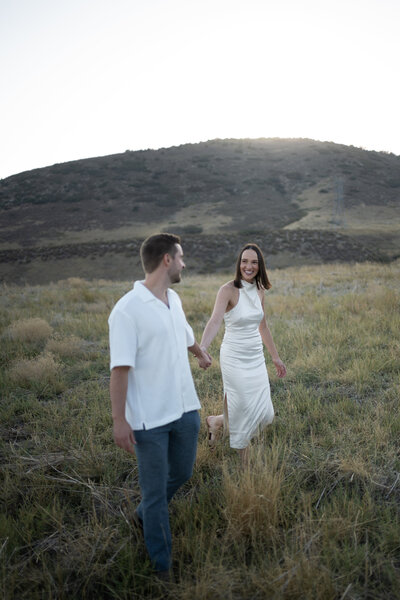 A couple walks hand in hand through a golden grassy field at sunset, surrounded by rolling hills. The woman, wearing a sleek white halter dress with a thigh slit, smiles warmly at her partner. Her dark hair is neatly styled, and she moves gracefully through the tall grass in strappy sandals. The man, dressed in a casual white short-sleeved button-up and dark jeans, gazes back at her with admiration. The soft, fading sunlight casts a dreamy glow over the landscape, capturing a moment of effortless romance and joy.