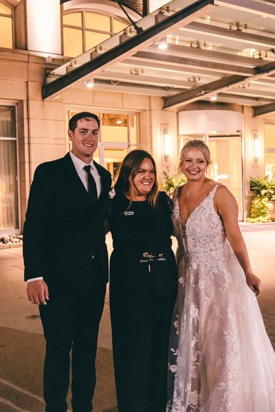 A bride in a lace gown, a groom in a suit, and a quad cities wedding planner in a black outfit smiling together outside a building at night.