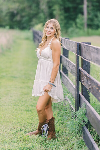 A senior girl in a white dress and brown boots smiling softly beside a rustic fence — Portrait photographer in Raleigh.